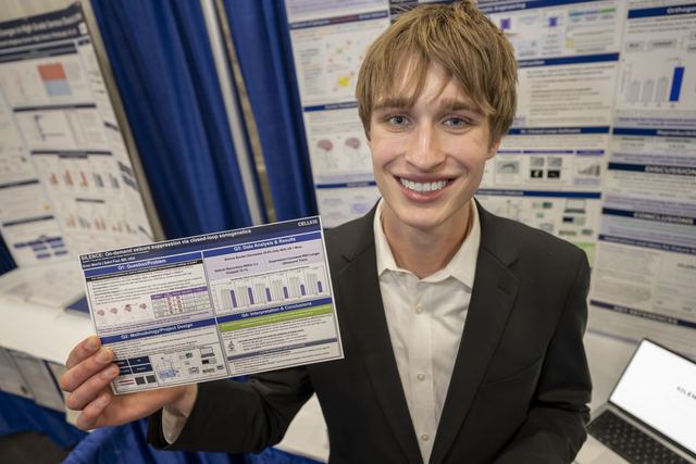 Official ISEF photo of Evan smiling and holding a printed mini-poster handout of his SILENCE project in front of his poster board, with a laptop visible at the edge of the booth