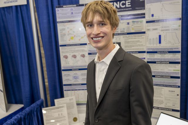 Official ISEF photo of Evan in a suit smiling in front of his SILENCE poster board, with the project title and a Regeneron banner visible at the left edge