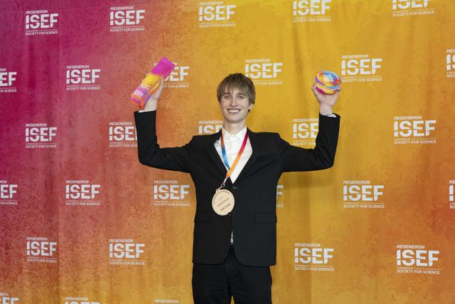 Celebratory backstage shot of Evan with both arms raised, holding his awards overhead, first-place medal around his neck, against the ISEF backdrop