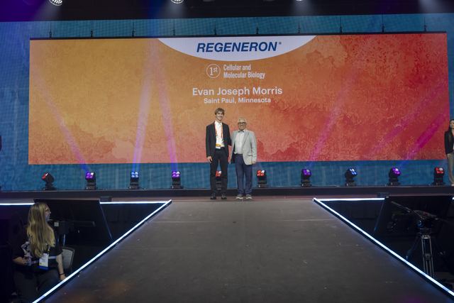 ISEF grand awards stage with the giant screen reading 'Regeneron, 1st, Cellular and Molecular Biology, Evan Joseph Morris, Saint Paul, Minnesota' while Evan stands with the presenter on the runway stage