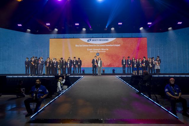 Wide shot of the ISEF awards stage with the screen reading 'Mary Sue Coleman Award for Life Science Innovation & Impact, Evan Joseph Morris, Saint Paul, Minnesota'; Evan stands center stage receiving the award with rows of other finalists lined up on stage