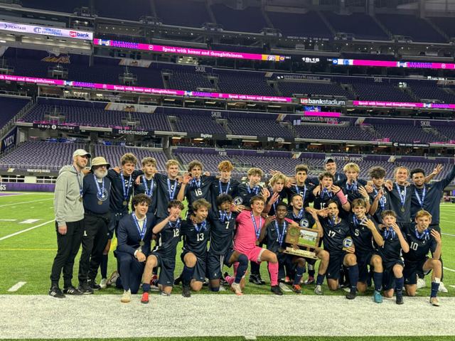 SPA boys soccer team posing with the state championship trophy on the field at U.S. Bank Stadium after the state three-peat; about 25 players and coaches with medals, stadium seating behind