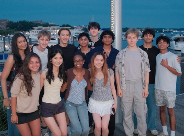 Evan with a large group of friends on a waterfront boardwalk at dusk, marina boats and city lights across the water behind them
