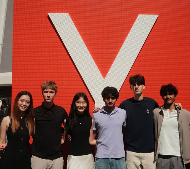 Evan, second from left, with five other students in bright sunlight in front of the Y Combinator logo on a red LED wall