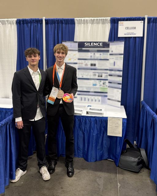 Evan Morris wearing his Regeneron ISEF award medallion beside a fellow finalist in front of the SILENCE poster at booth CELL038