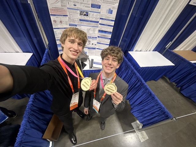 Wide-angle selfie of Evan and a friend wearing gold first- and second-place medals in front of a science fair poster booth with orange lanyards, likely a state or regional fair preceding ISEF