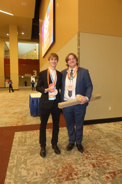 Evan and another male finalist in suits standing in a convention center lobby near a 'Hall 3' entrance with ISEF signage on a screen above; Evan holds a round rainbow ISEF award disc and the other student holds a medal and a long box