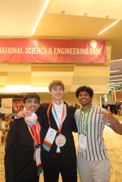 Evan with arms around two male friends under the ISEF banner; one holds up a silver medal, Evan wears a badge with a 'First Place' ribbon