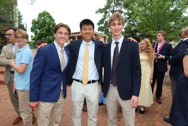 Evan and two friends in navy blazers and ties posing outdoors on brick pavers at a dressy school event; crowd in background including a student with a lei, suggesting graduation or an end-of-year ceremony