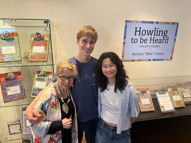 Evan and a teenage girl with an older woman in front of a gallery exhibit sign reading 'Howling to be Heard, Epilepsy Exhibit, Barbara "Babs" Larson', surrounded by display cases of decorated art books
