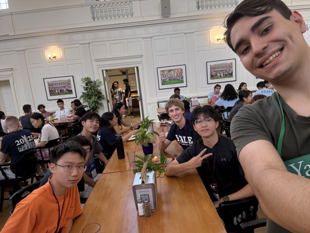 Evan giving a thumbs-up in a group selfie at a long communal table in a bright, wood-paneled Yale dining hall during a summer program