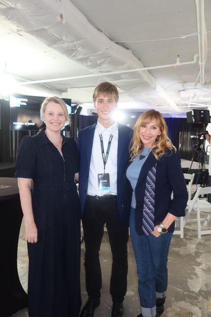Flash photo of Evan in a navy blazer with an event lanyard flanked by two adult women in a backstage/green-room area with TV cameras and a branded backdrop