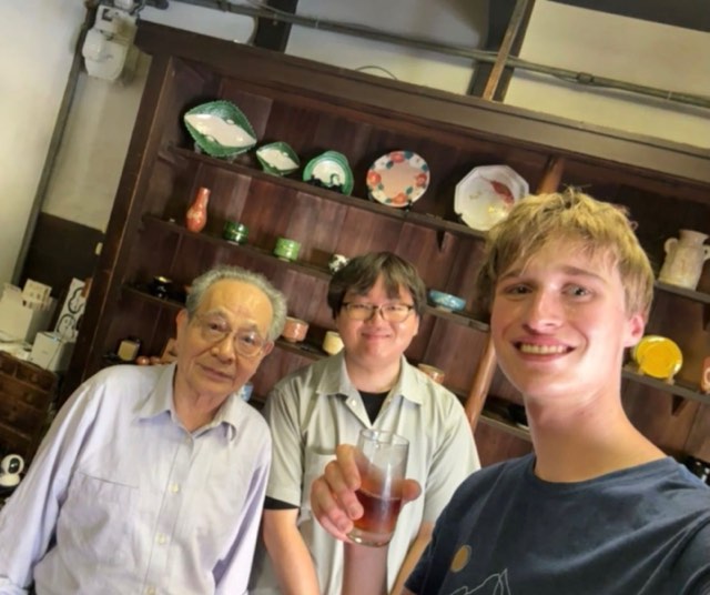 Evan raising a glass in a selfie with two hosts in front of tall wooden shelves of glazed Japanese ceramics