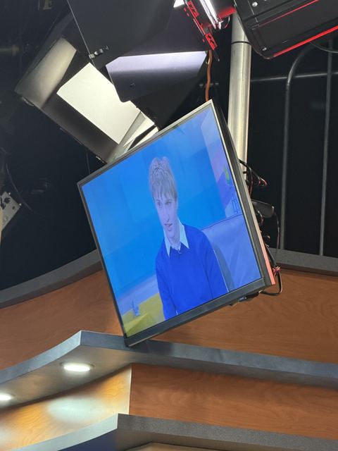 Tilted studio monitor suspended in the TV set showing Evan live on air in a blue sweater against the show's blue backdrop; studio lights and rigging around it