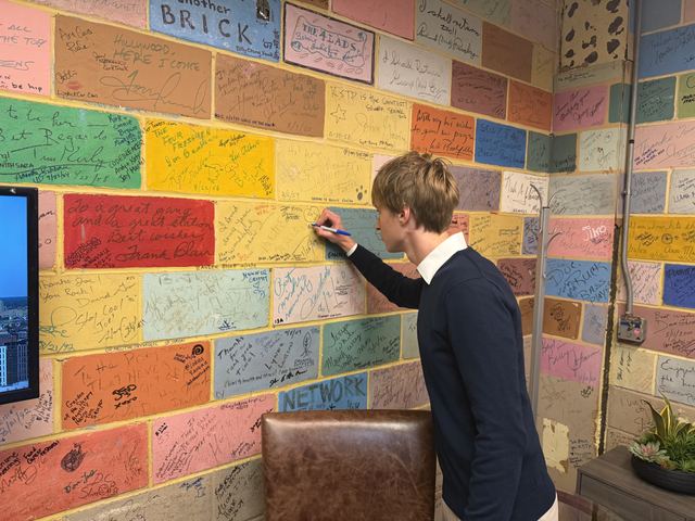 Evan, seen from behind in a navy sweater, signs a pastel-painted signature brick wall covered with guest autographs at the TV station