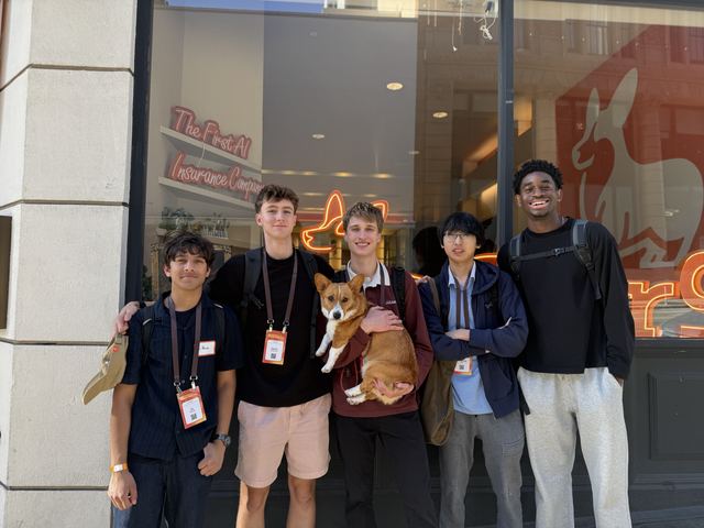 Five teens, one holding a corgi, pose outside a storefront with a neon sign reading 'The First AI Insurance Company', looks like a San Francisco startup-tour or travel stop