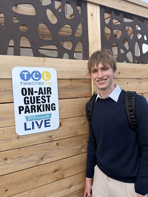 Evan smiling next to the 'TCL Twin Cities Live / Minnesota Live On-Air Guest Parking' sign on a wooden fence, wearing a navy sweater and backpack