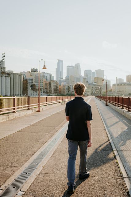 Striking professional shot of Evan from behind, walking down the Stone Arch Bridge toward the Minneapolis skyline in golden-hour light, strong sense of place and forward motion