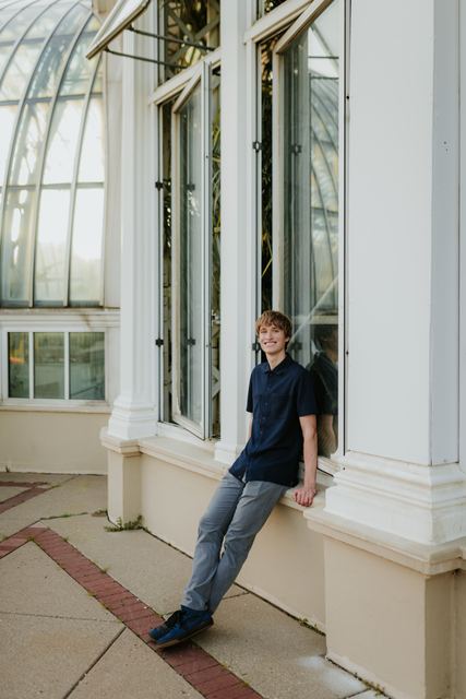 Same conservatory-window pose as IMG_4106 but smiling at the camera, the stronger frame of the pair; navy shirt against white columns and glass dome