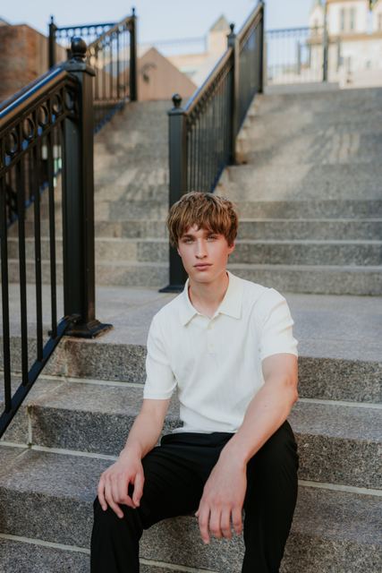 Evan in a cream polo and black pants seated on wide granite steps with black iron railings in a downtown setting, clean urban editorial portrait