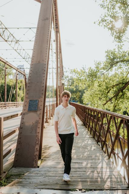 Evan walking across a historic iron truss bridge over a river with sun flaring through trees, dynamic, atmospheric professional shot with strong leading lines