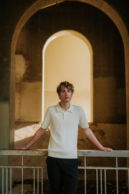 Moody professional portrait under a concrete arched underpass, Evan in a cream knit polo leaning back against a railing with arms outstretched, warm light glowing through the arch behind him