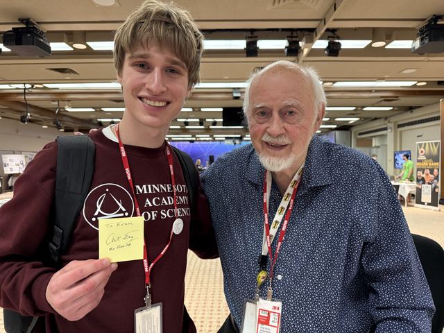 Evan in a Minnesota Academy of Science sweatshirt posing with an elderly man wearing a 3M lanyard at an indoor science fair venue; Evan holds a yellow Post-it note signed 'To Evan' by the man, who appears to be Art Fry, the Post-it Note inventor