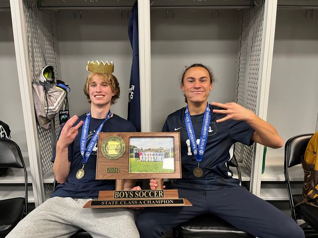 Locker-room celebration: Evan wearing a gold paper crown and a state-championship medal sits with a teammate, jointly holding a 'Boys Soccer State Class A Champion 2024' plaque trophy