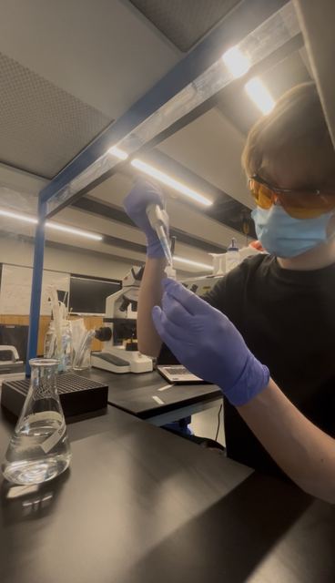 Evan in orange safety glasses, face mask, and purple nitrile gloves loading a micropipette under bright bench lighting, with an Erlenmeyer flask and microscope nearby; strong action shot of wet-lab work