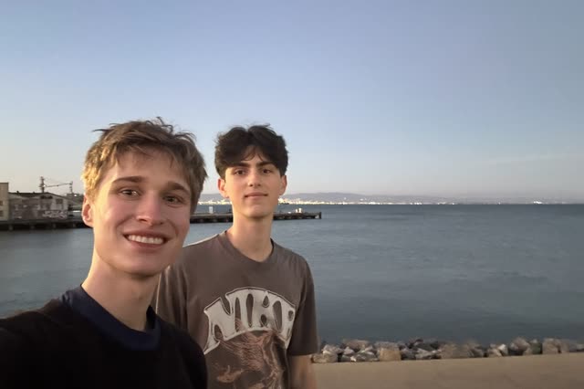 Evan and a friend in a waterfront selfie at dusk, a pier and city lights across the bay behind them