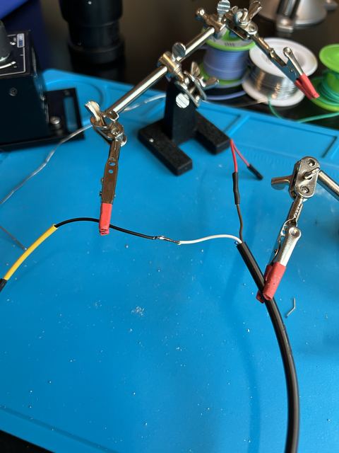 Close-up of a soldering workbench: helping-hands clamps holding wires mid-solder over a blue silicone mat, with solder spools and equipment in the background