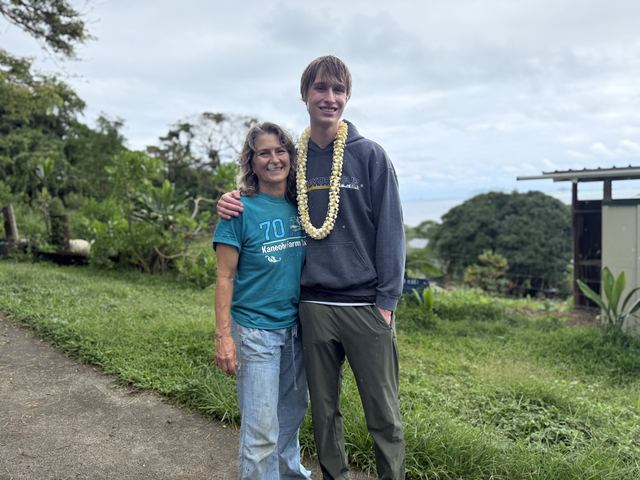 Evan wearing a white flower lei stands smiling with his arm around an older woman in a 'Kaneohe Farm' t-shirt, on a farm hillside with the ocean and greenhouse structures behind
