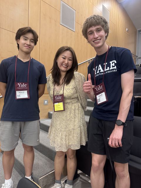 Evan (right, thumbs up) with two other teens in a lecture hall, all wearing Yale Young Global Scholars lanyards and name badges