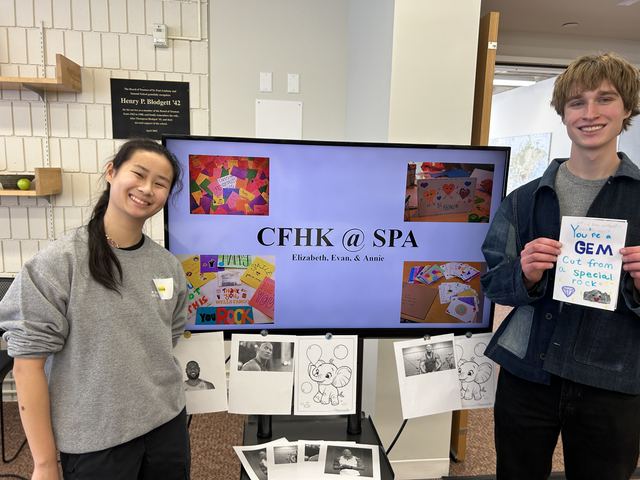 Evan and a fellow student flank a TV displaying a 'CFHK @ SPA' slide (Cards for Hospitalized Kids at St. Paul Academy) credited to 'Elizabeth, Evan, & Annie'; Evan holds up a handmade card reading 'You're a GEM cut from a special rock'