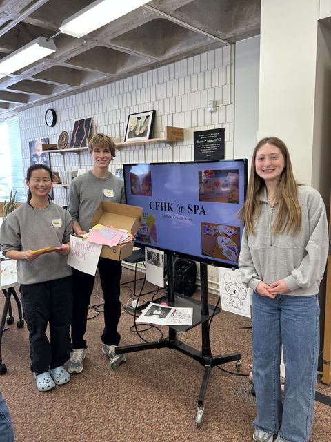 Wider shot of the CFHK @ SPA card-making event: Evan holds a box of finished cards, flanked by two other students, with the presentation screen, a 'Finished Cards' sign, and coloring pages around a classroom/library space