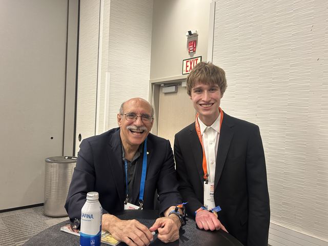 Evan in a dark suit with an orange ISEF lanyard poses at a small table with a smiling older scientist in a blazer (mustache, glasses, lapel pin), reads as a meet-and-greet with a distinguished speaker or Nobel laureate at ISEF 2026