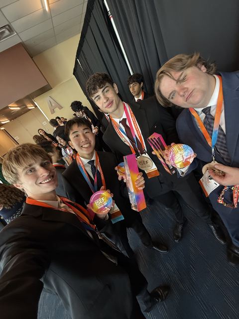 Celebratory selfie of Evan with three other finalists in suits, wearing ISEF medals and lanyards and holding colorful ISEF award trophies backstage by dark curtains, clearly the grand awards celebration