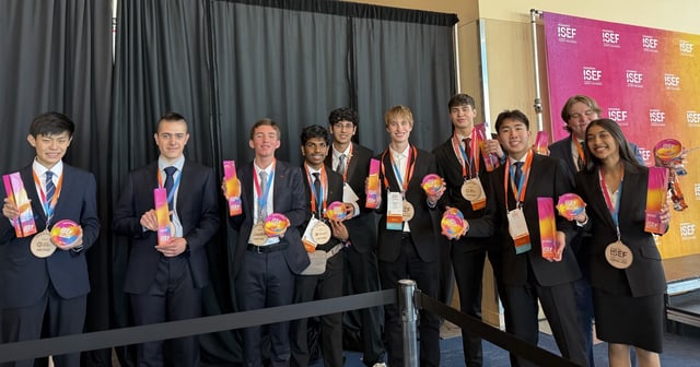Evan, center, with other Regeneron ISEF finalists in suits holding trophies and rainbow award discs in front of the ISEF backdrop in the convention hall