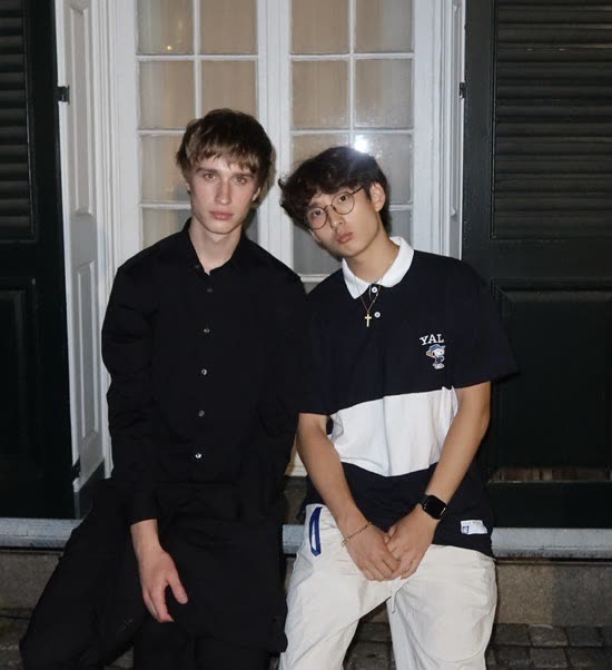 Evan Morris in a black shirt sitting beside a friend in front of a white-framed door at night