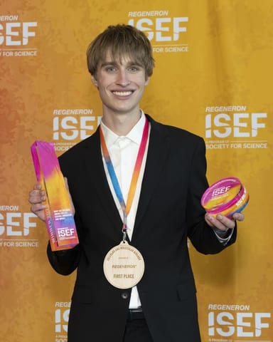 Evan Morris backstage at Regeneron ISEF 2026, wearing his wooden first-place medal and holding his Mary Sue Coleman Award trophy and award disc