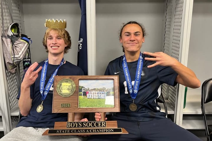 Evan Morris in a gold paper crown and a championship medal, sitting beside a teammate with the state championship plaque